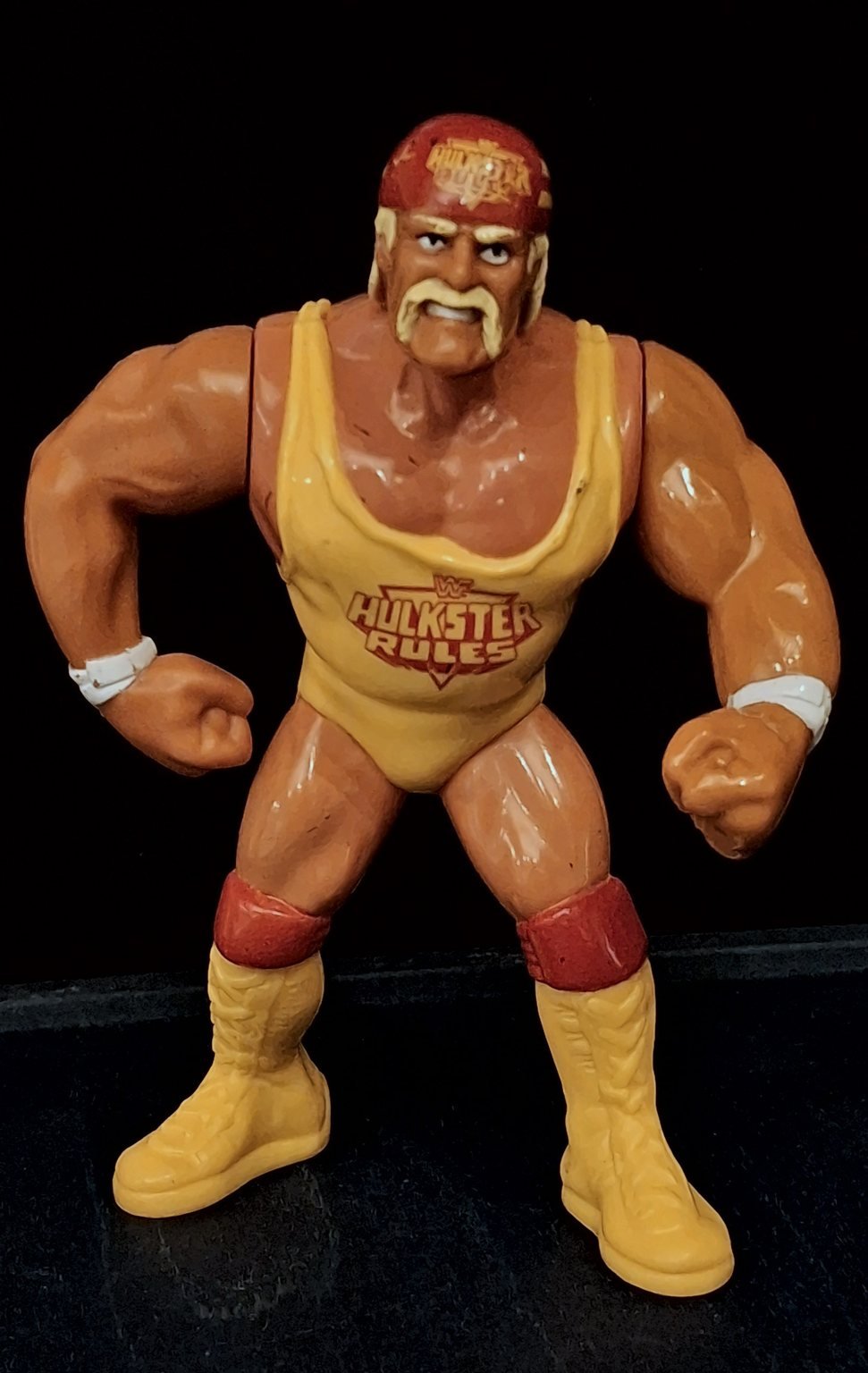 Hulk Hogan in piedi, di fronte, con la bandana rossa, la canotta gialla “Hulkster Rules”, le braccia piegate e i pugni chiusi