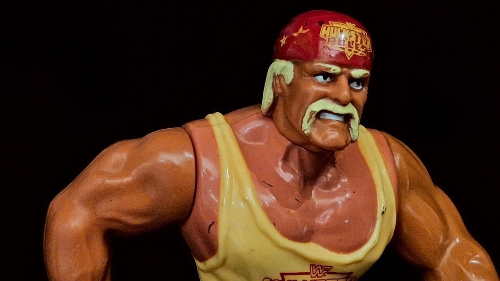 Il volto di Hulk Hogan girato verso la sua sinistra, con i baffi biondi a ferro di cavallo e i denti scoperti