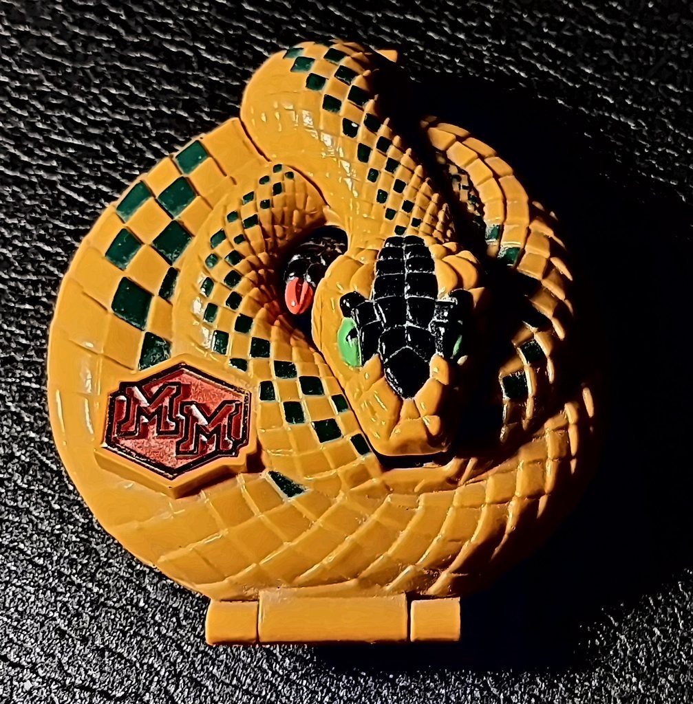 Il guscio chiuso visto dall'alto: un serpente arrotolato color ocra con i rombi verdi sul dorso, la testa al centro e il distintivo rosso “MM” sul fianco